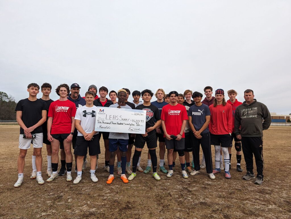 A soccer team poses outdoors, holding a large donation check for LEHS Soccer.