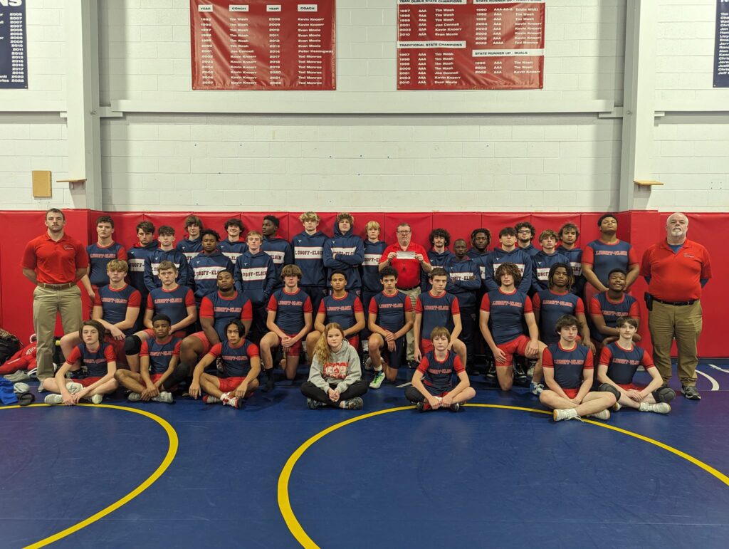 Group photo of a high school wrestling team and coaches in matching uniforms on a blue mat in a gym.