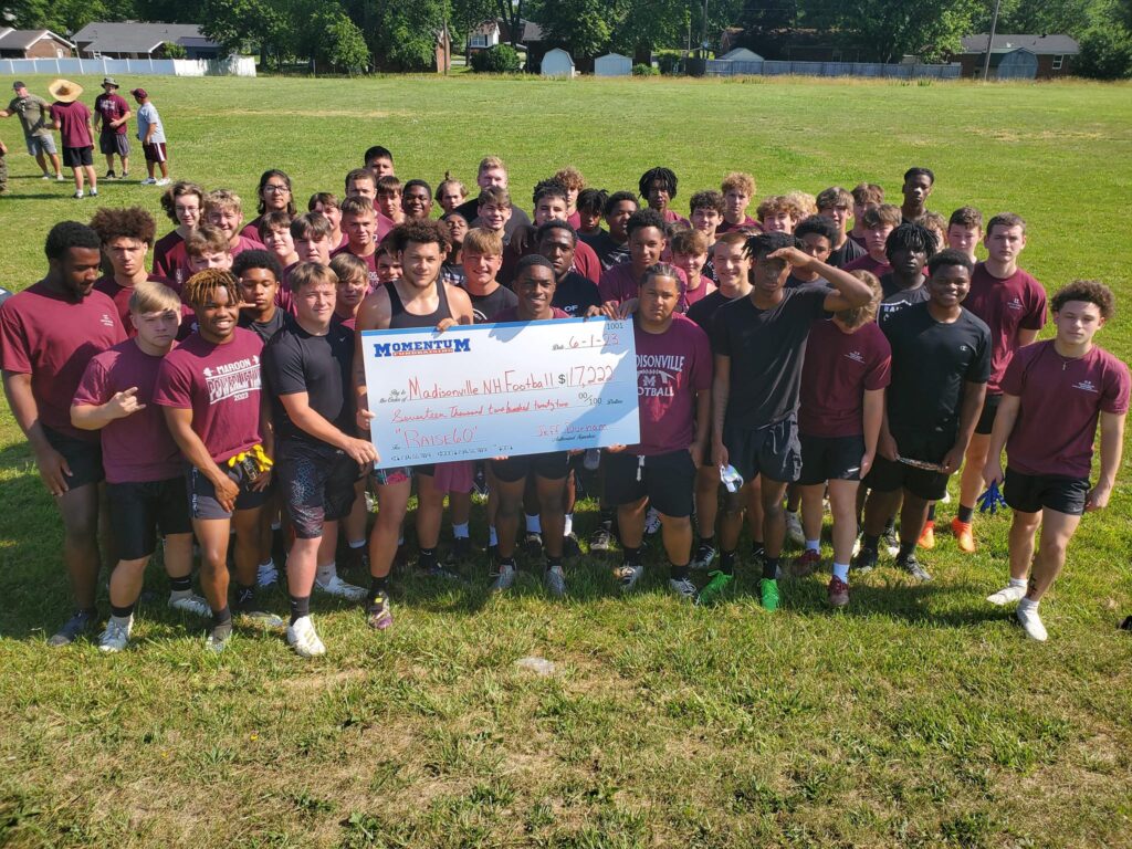 A large group of teens in maroon shirts hold a giant check, standing together on a grassy field.