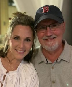 Smiling woman and man in a cap pose together indoors for a close-up photo.