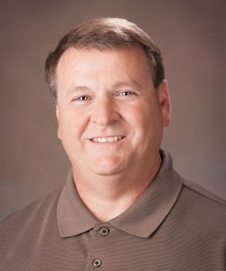 Smiling middle-aged man with short brown hair wearing a brown polo shirt, posing in front of a plain background.